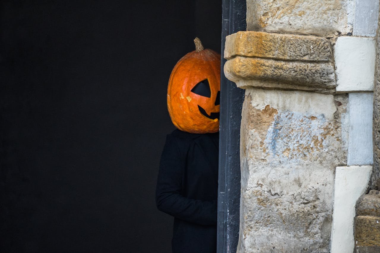 A person wearing a pumpkin mask peeks from behind a stone wall.